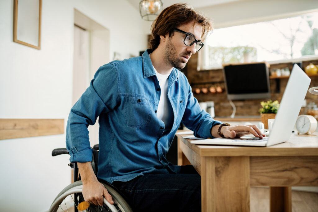 Man met bril, in rolstoel, werkt op een laptop.