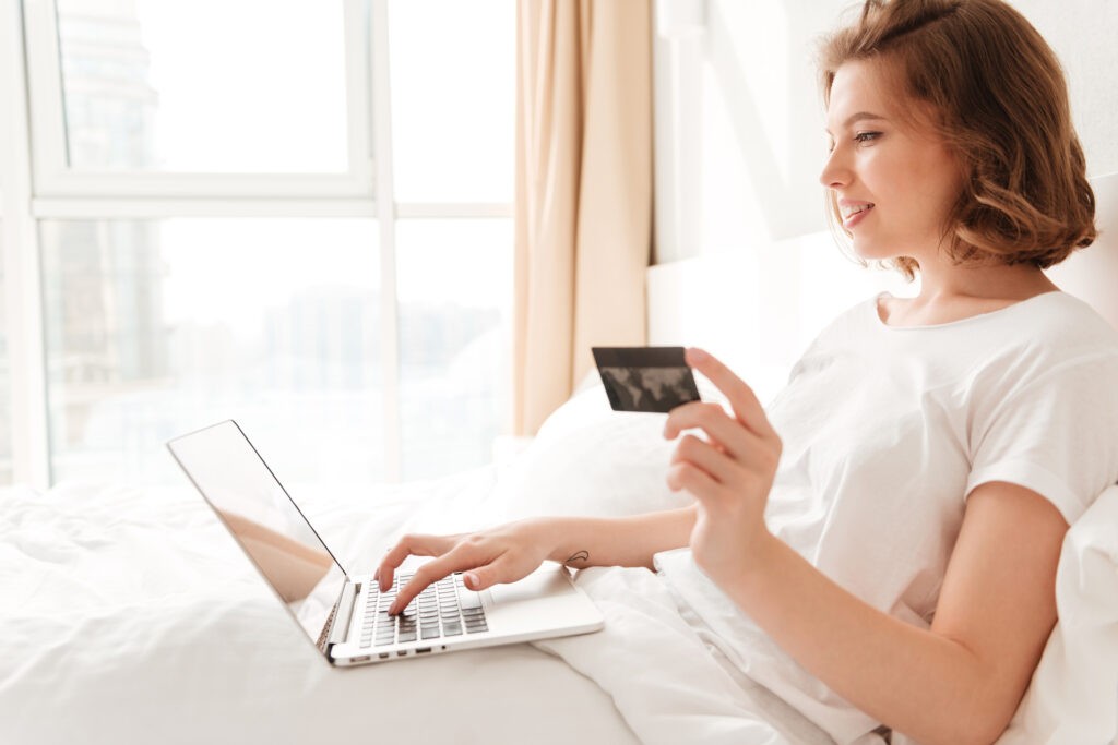 Vrouw zittend in bed met laptop op schoot en een creditcard in haar hand
